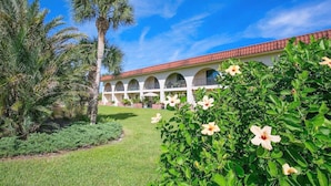 Un edificio con techo de tejas rojas, ventanas arqueadas y un patio con palmeras e hibiscos.