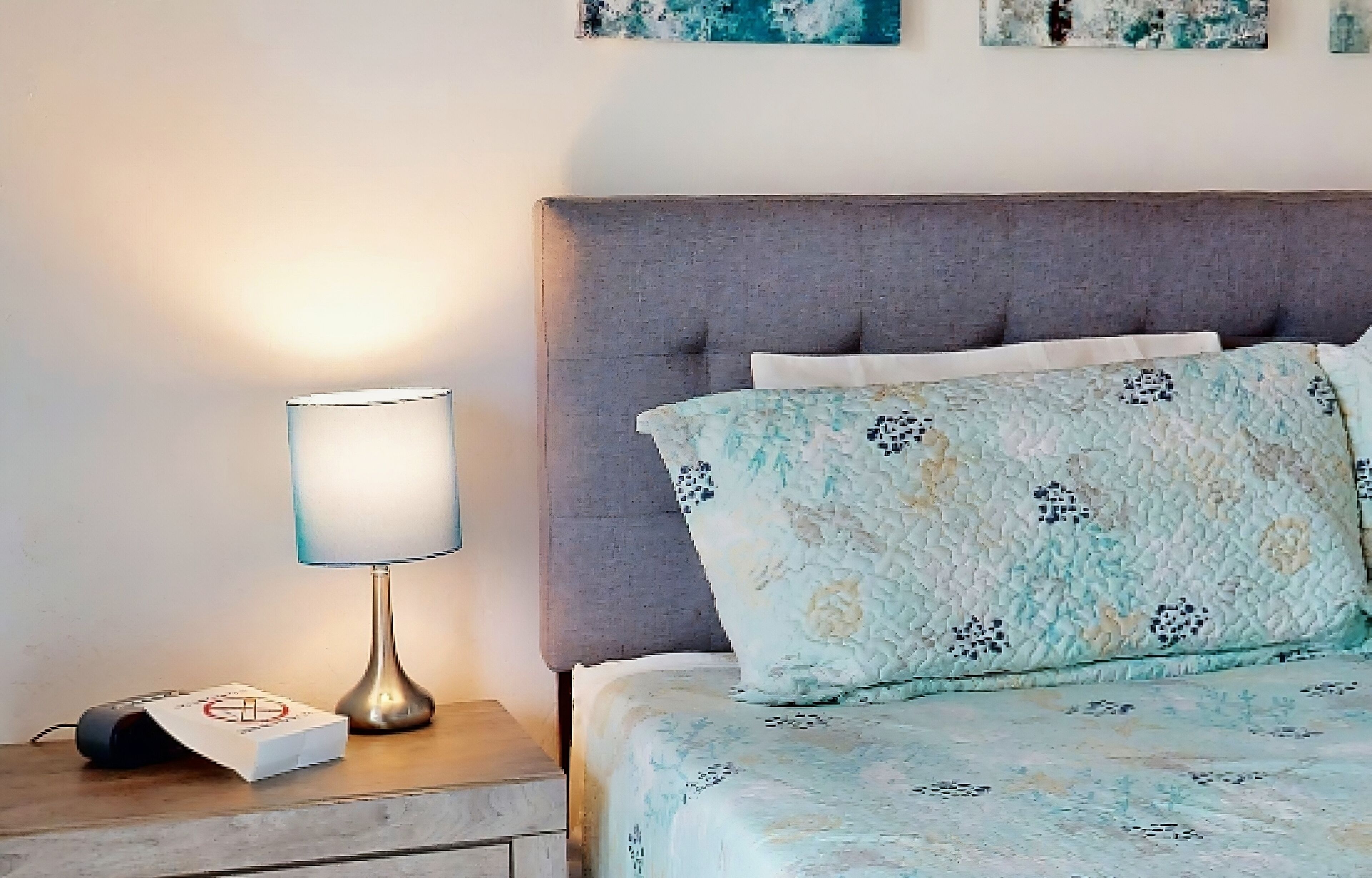 A neatly made bed with floral bedding, a bedside lamp, and a book on a wooden nightstand.