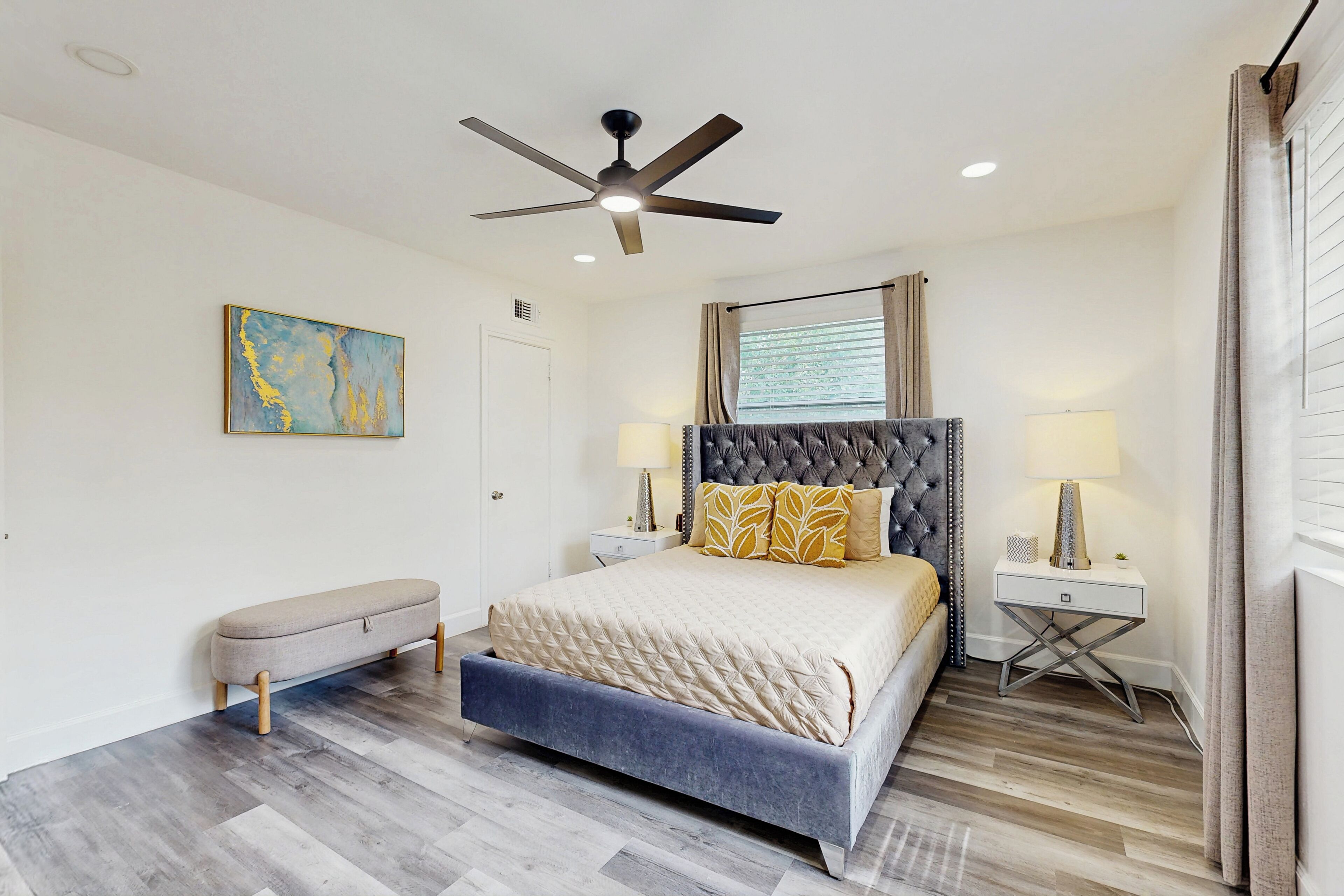A bedroom with a bed, a bench, two bedside tables, a ceiling fan, and a window with curtains.