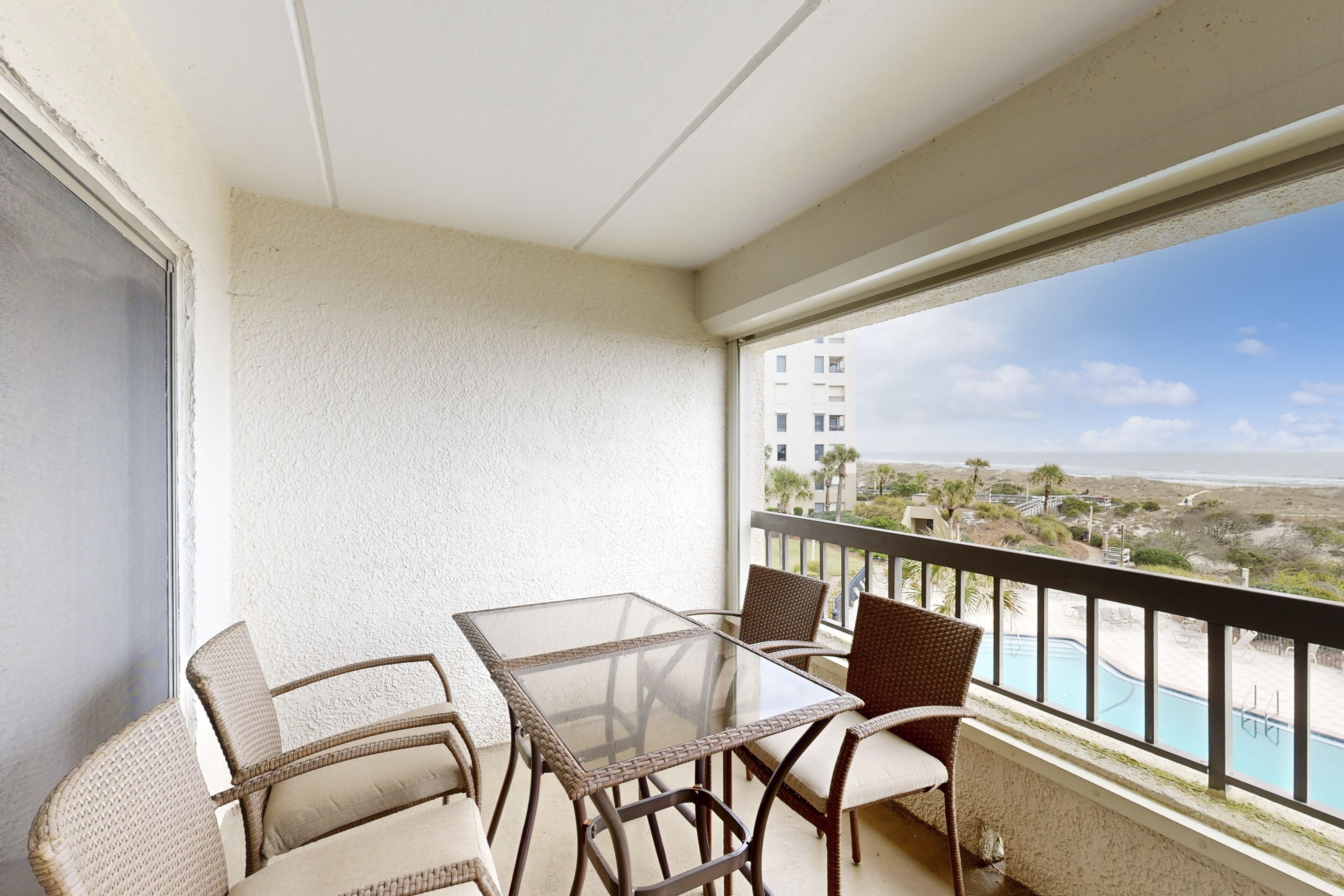 A balcony with a glass table and wicker chairs, overlooking a pool and a beach.