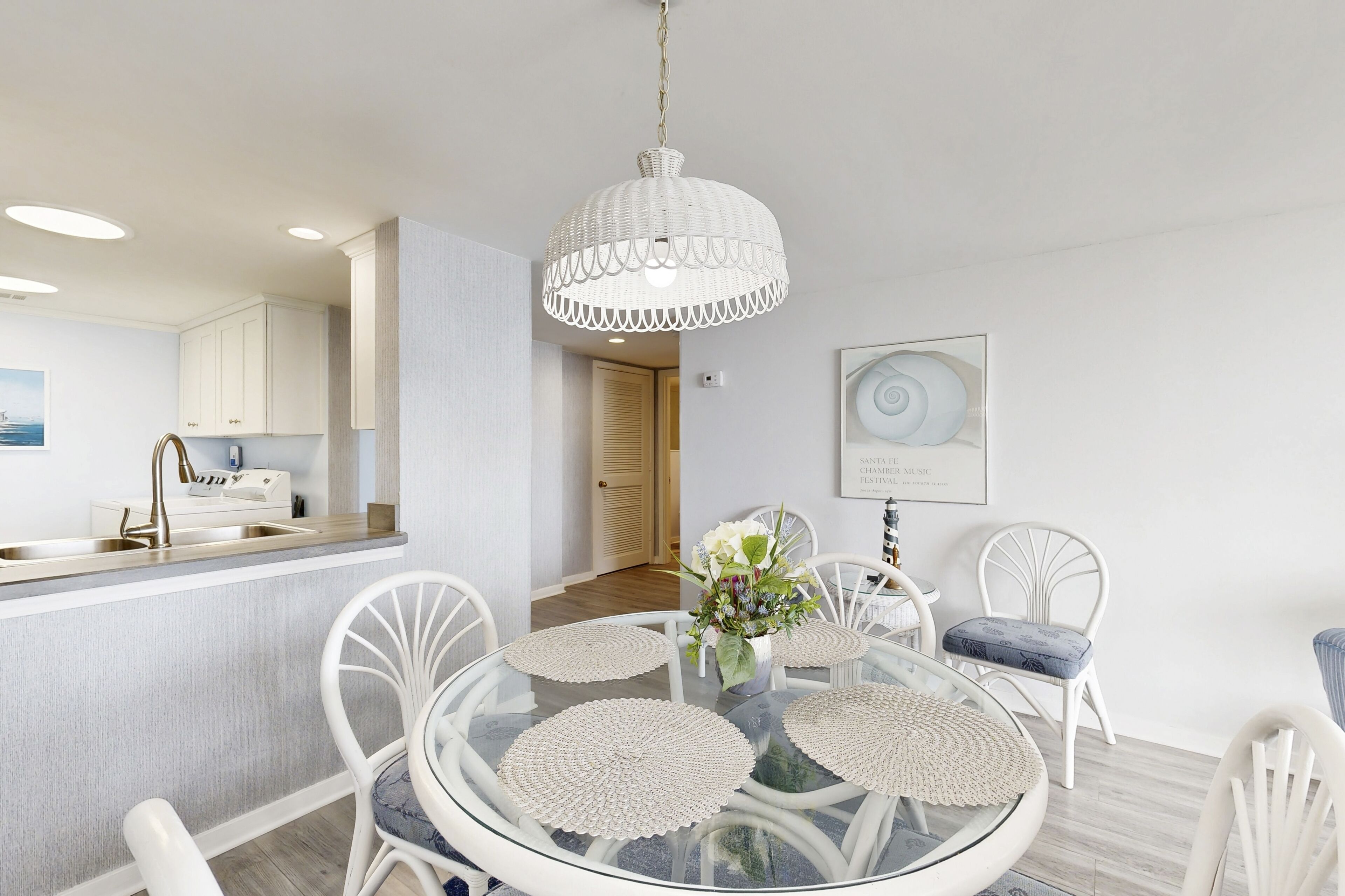 Un comedor con una mesa redonda de vidrio, sillas blancas, una lámpara colgante blanca y una cocina al fondo.