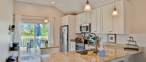 A modern kitchen with granite countertops, white cabinets, and stainless steel appliances. There is a balcony with a view of greenery.