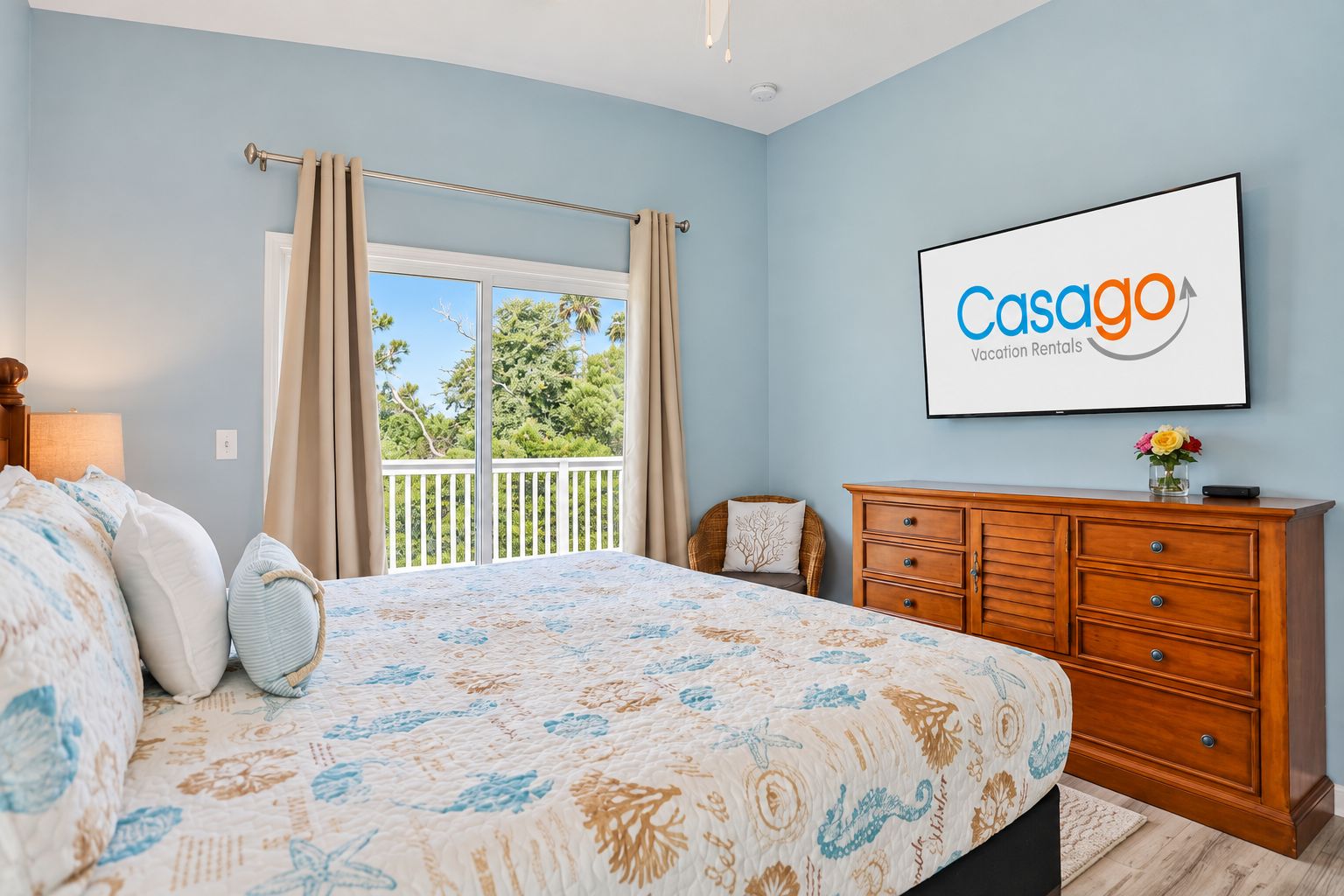 A bedroom with a bed, a dresser, a television displaying 'Casago Vacation Rentals,' and a balcony with curtains.
