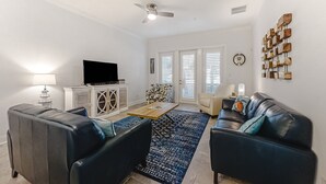 A living room with a sofa, a coffee table, a TV, and a ceiling fan. - Updated Townhome Near the Ocean W/private Patio, Shared Pool, Central A/C (Fernandina Beach)
