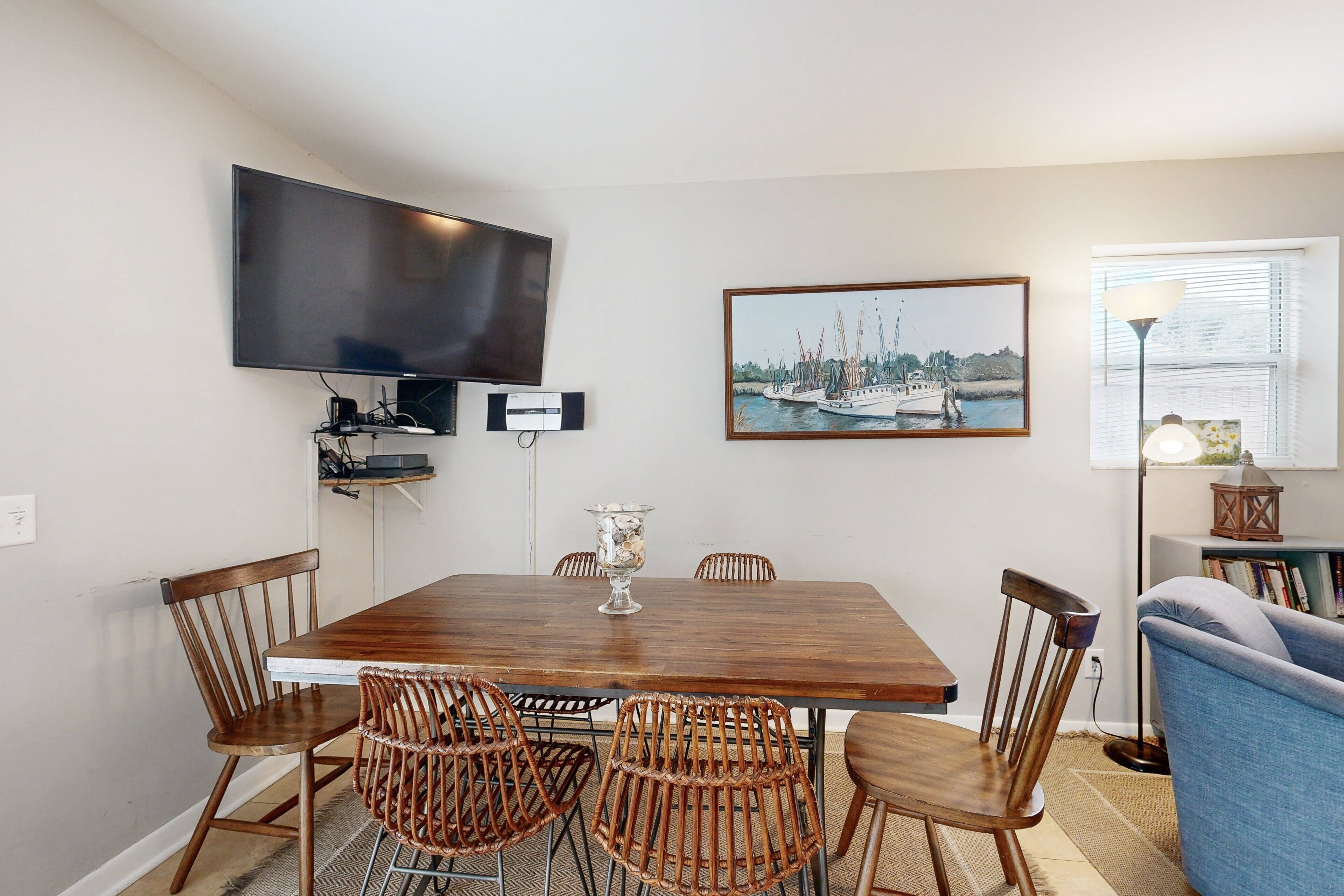 A dining area with a wooden table, wicker chairs, a flat-screen TV mounted on the wall, and a framed painting above the table.