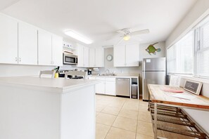 A modern kitchen with white cabinets, a central island, a microwave, a refrigerator, and a ceiling fan. - Sparkling Retreat Steps to Beach Fenced Backyard Snowbird Availability (Fernandina Beach)