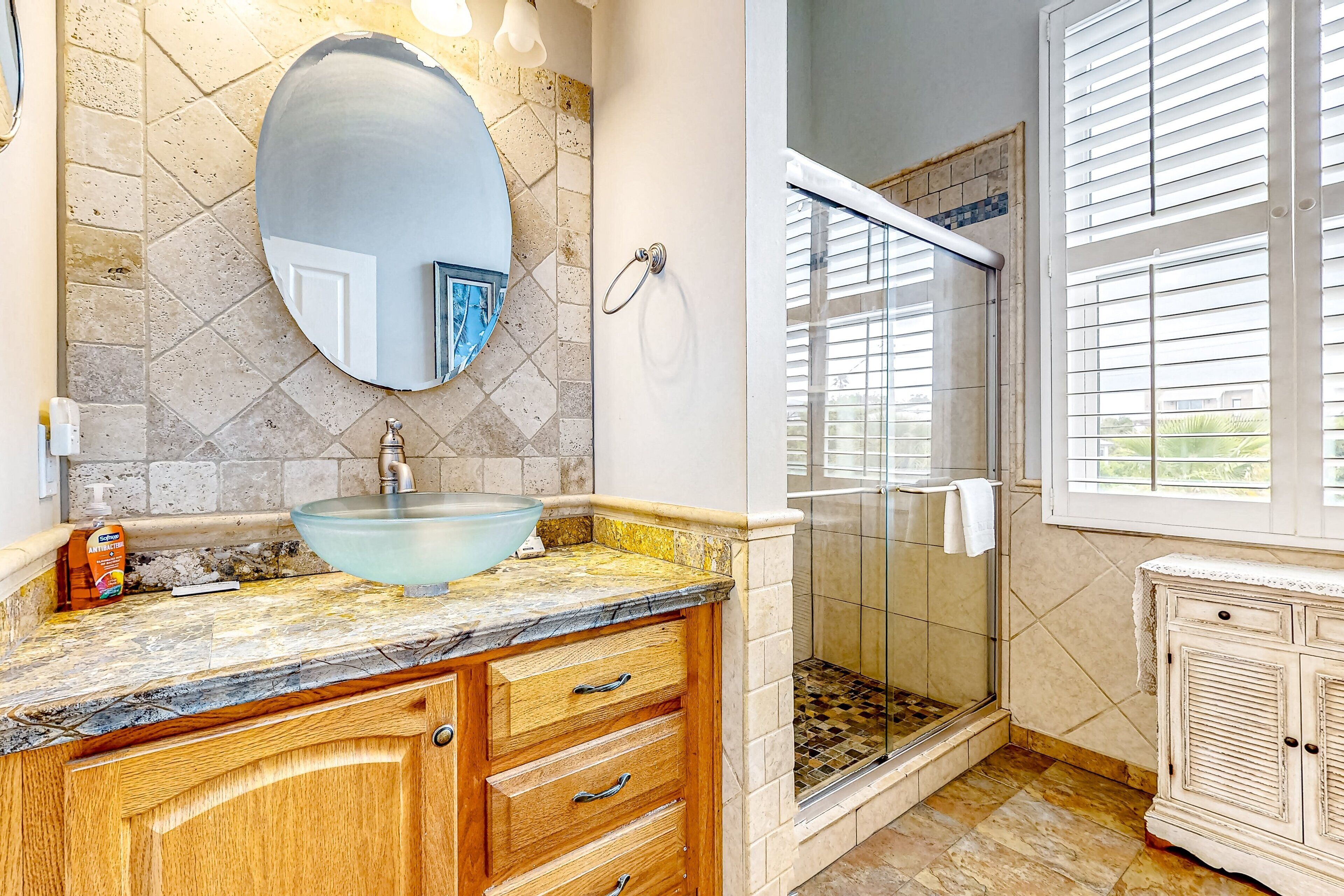 A bathroom with a glass sink, wooden vanity, a large mirror, a shower with glass doors, and a window with blinds.
