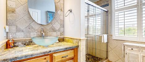 A bathroom with a glass sink, wooden vanity, a large mirror, a shower with glass doors, and a window with blinds.