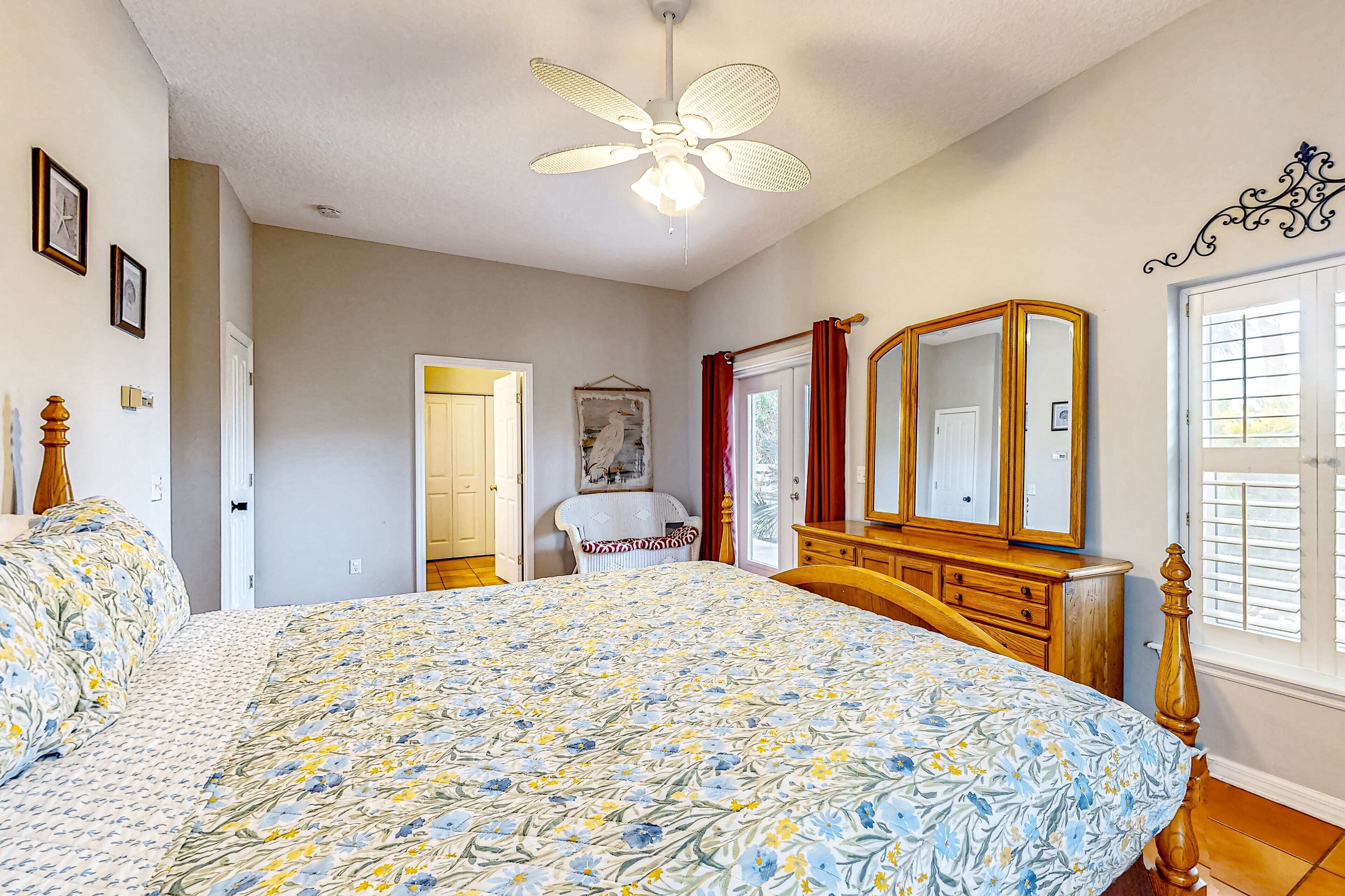 A bedroom with a large bed, a ceiling fan, a wooden dresser, a mirror, and a window with blinds.