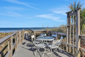 Eine Holzterrasse mit rundem Tisch und Stühlen, mit Blick auf einen Strand und den dahinterliegenden Ozean.