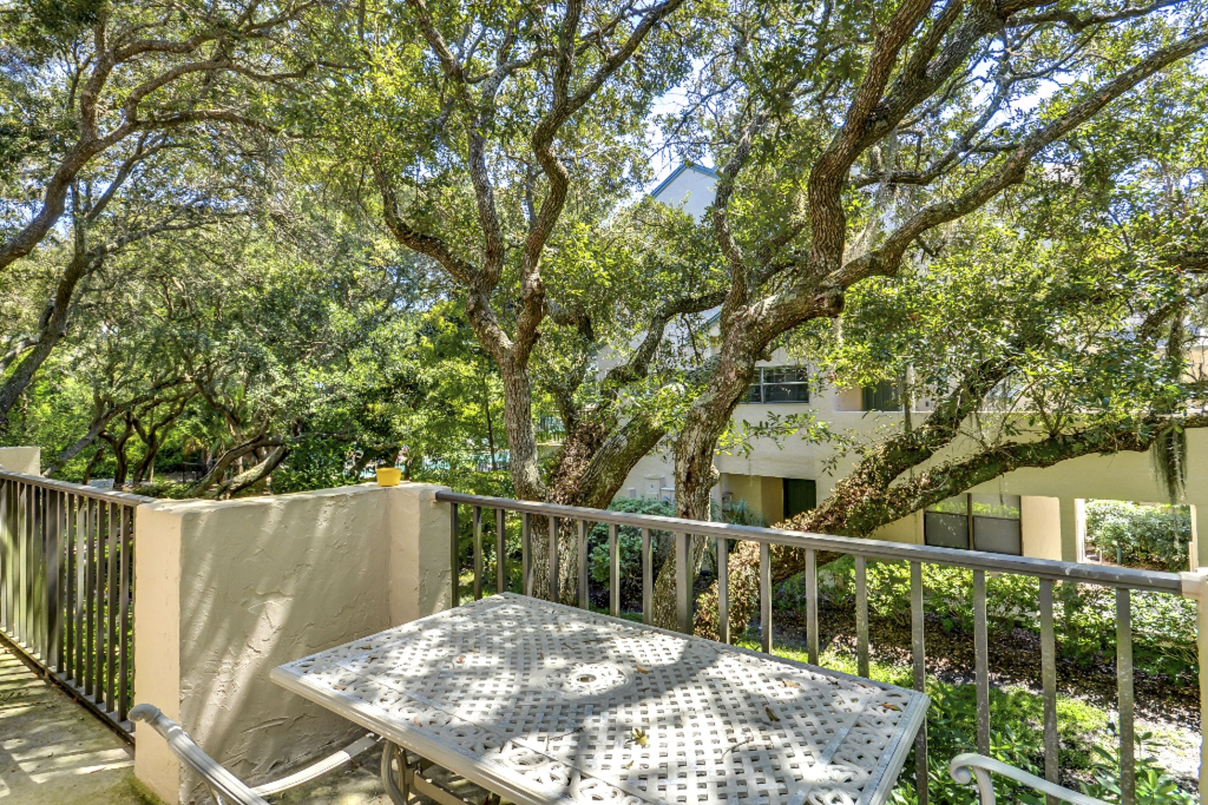 A balcony with a metal table and chairs, surrounded by trees and a building in the background.