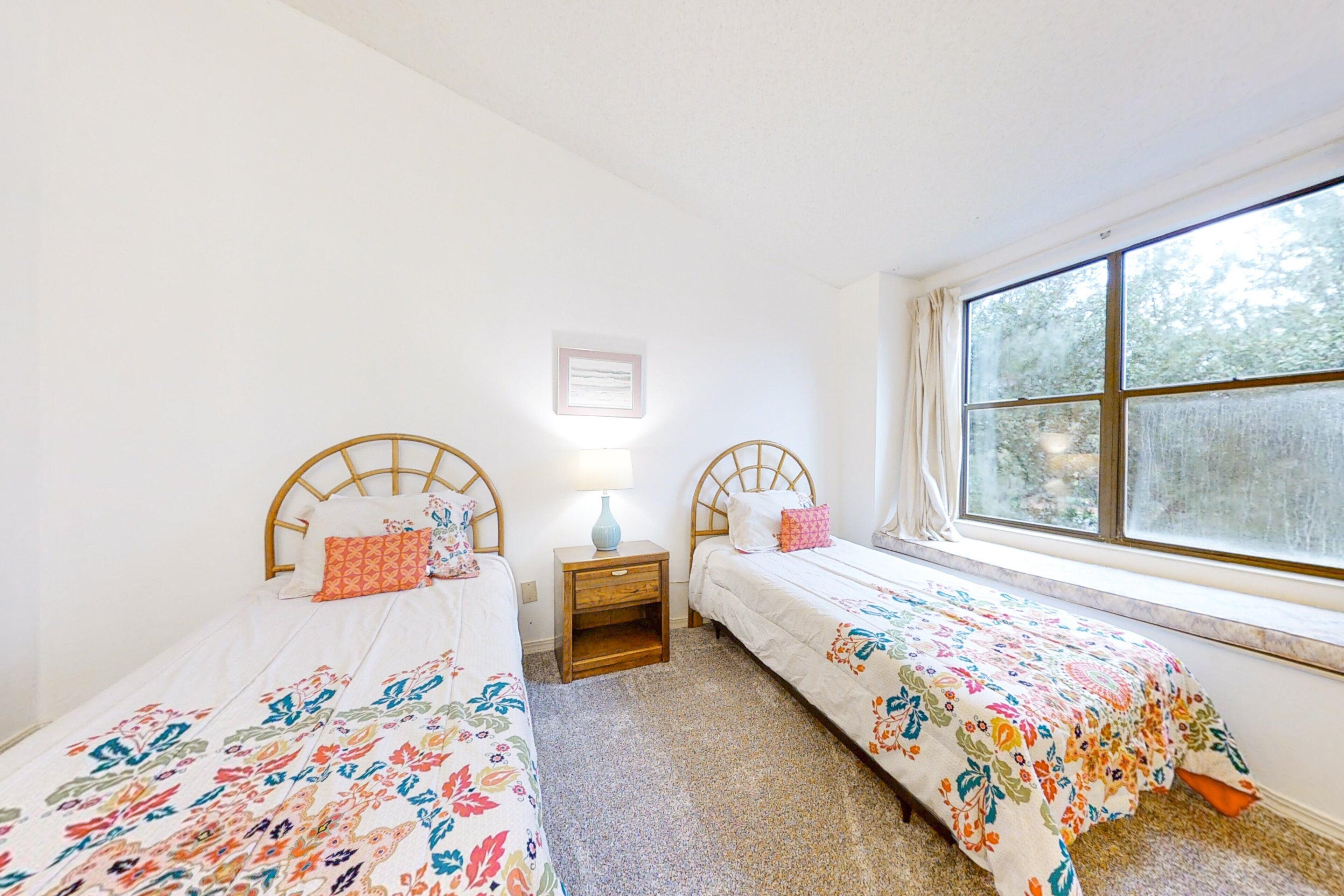 A bedroom with two single beds, a small bedside table with a lamp, a window with curtains, and a framed picture on the wall.