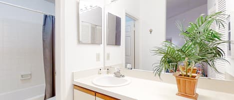 Un baño con lavamanos blanco, tocador de madera, un espejo y una planta en maceta.