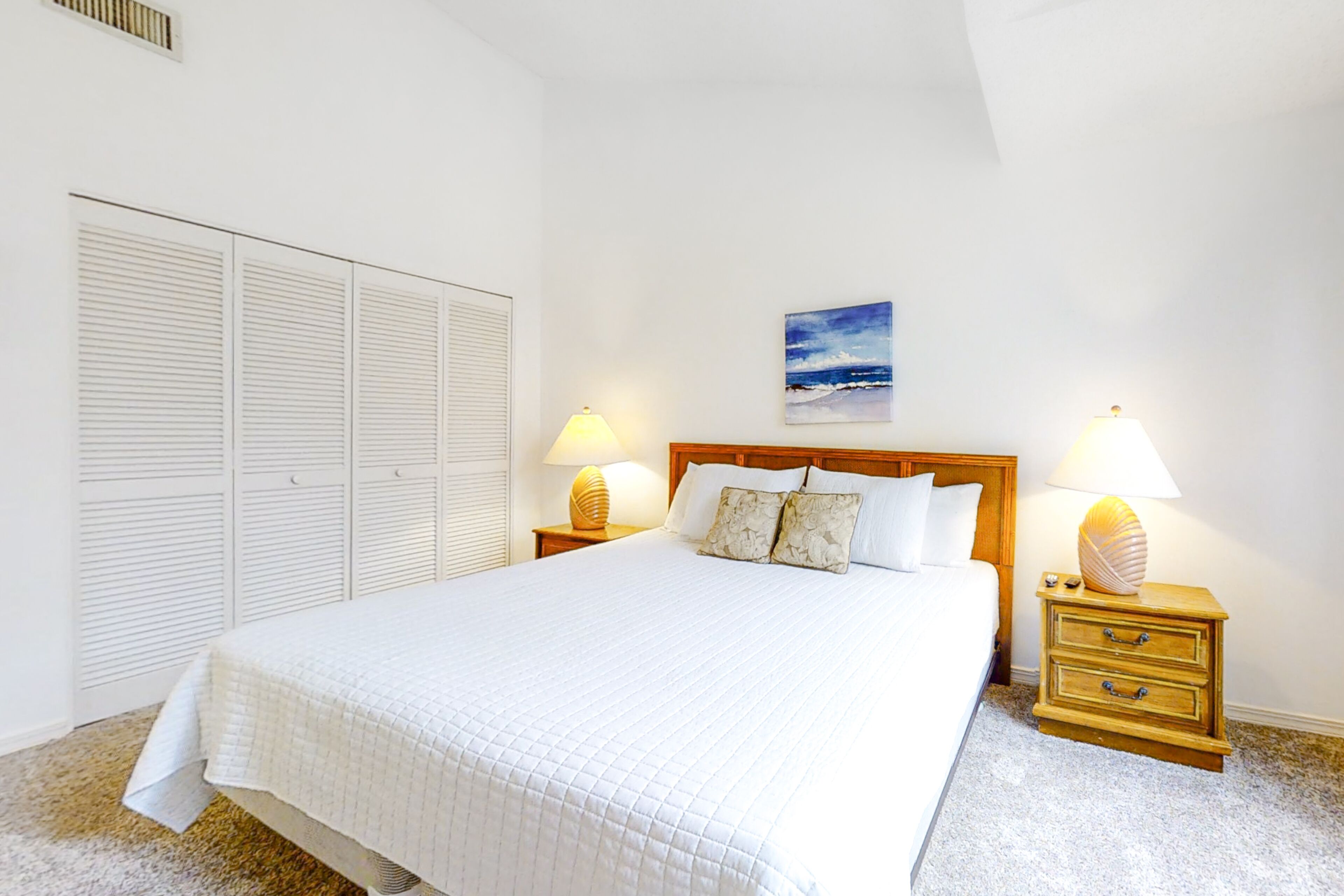 A neatly made bed with a quilted white comforter, two bedside lamps, a wooden nightstand with drawers, and a wall-mounted painting of a beach scene.