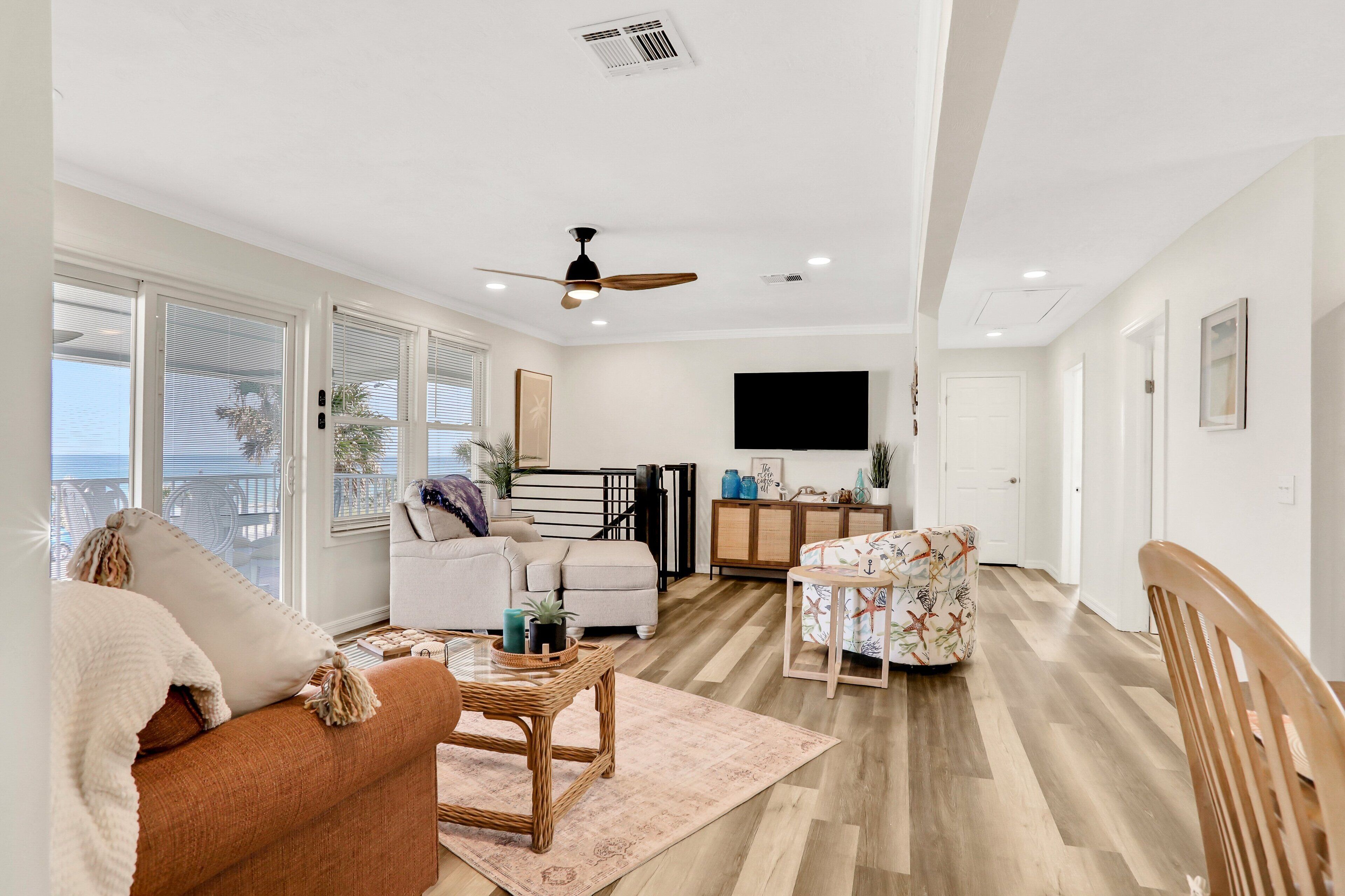 A spacious living room with a sofa, armchair, and coffee table. A ceiling fan and TV are mounted on the wall.