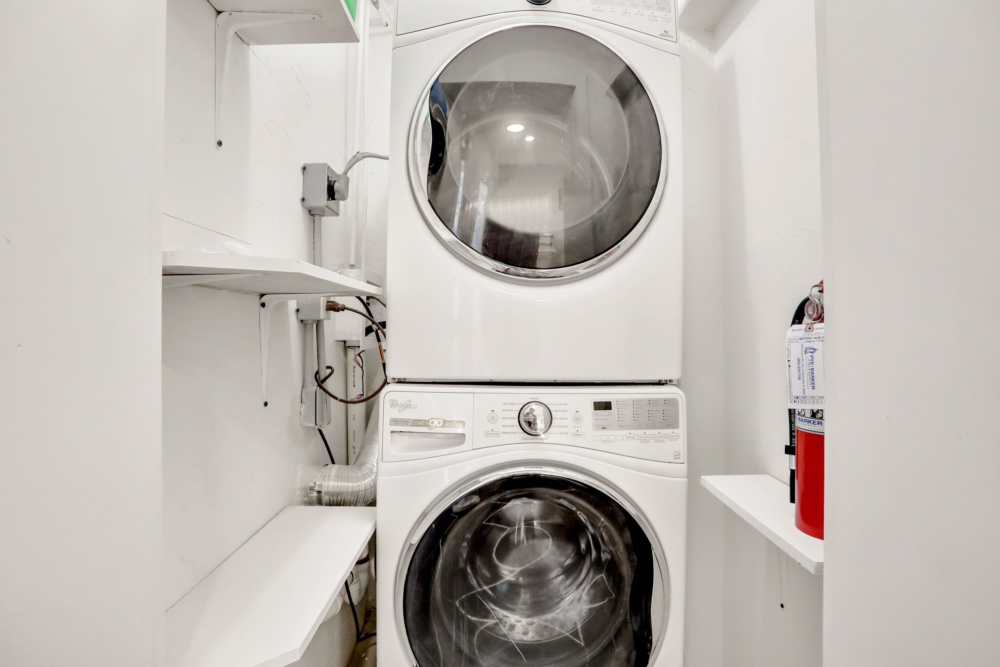 Een kleine wasruimte met een voorlader wasmachine en droger, een klein plankje en zichtbare leidingen.