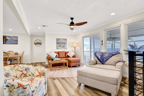 A spacious living room with a large window, a ceiling fan, and a dining area with wooden furniture. - Updated Home Across From Beach With Covered Balcony, Central AC, & Washer/dryer (Flagler Beach)