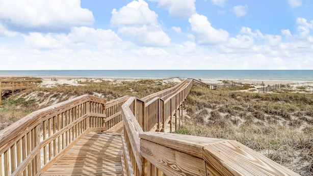 Pasarela de madera que conduce a una playa con dunas y cielo despejado.