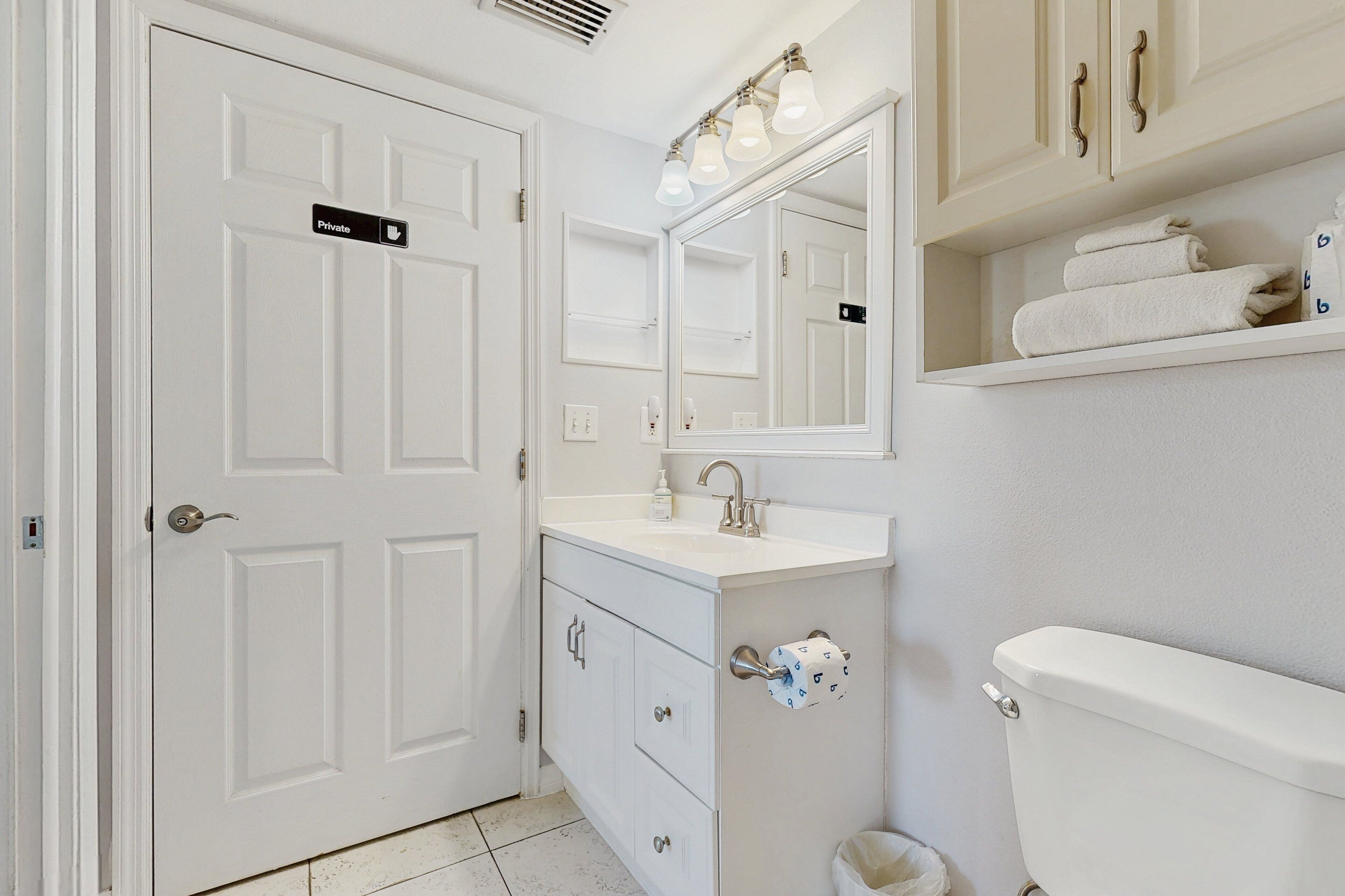A private bathroom with a white sink, mirror, and toilet. There are towels on a shelf and a trash can.