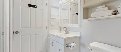 Baño privado con lavamanos blanco, espejo y retrete. Hay toallas en un estante y un cubo de basura.
