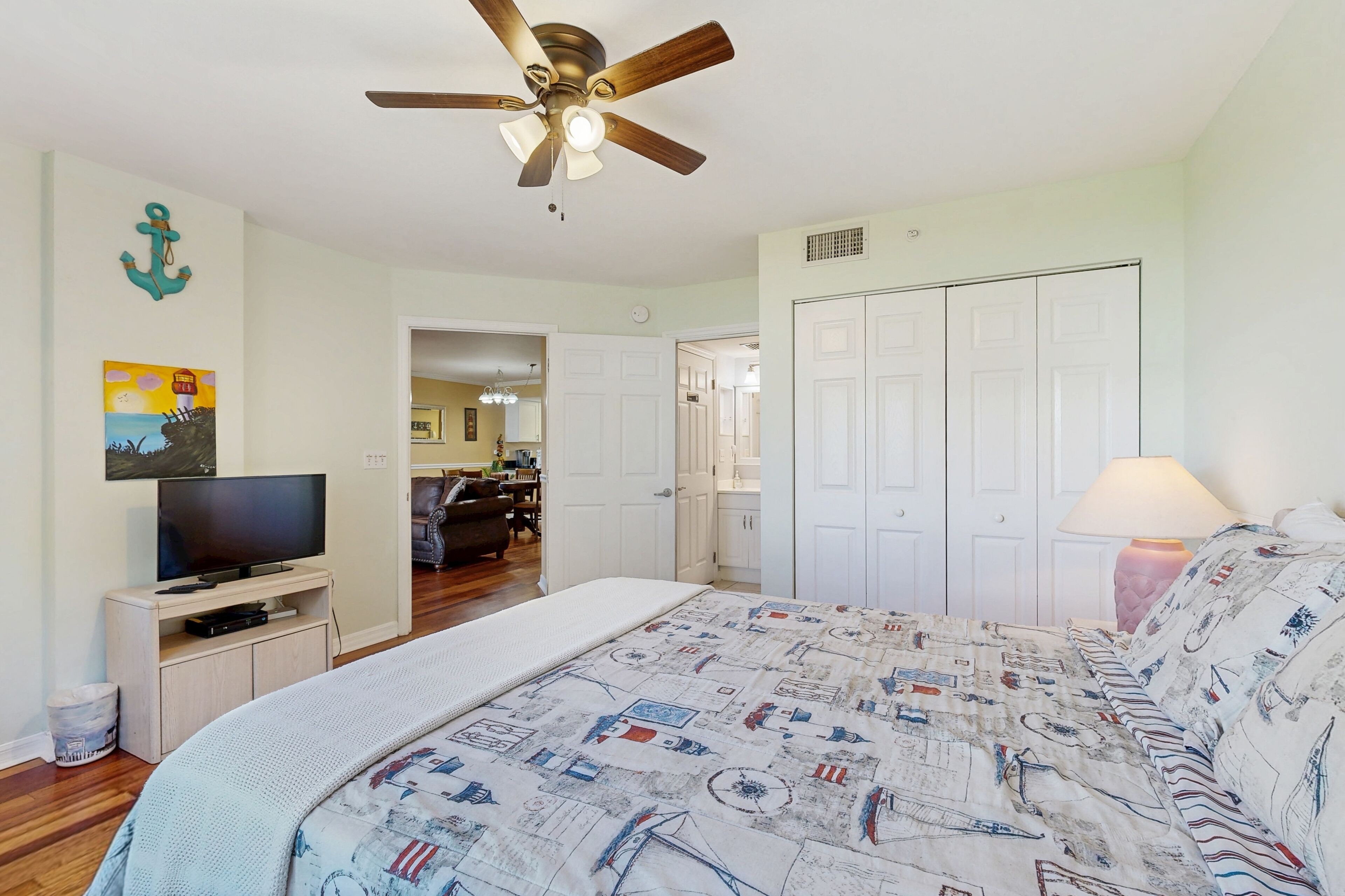 A bedroom with a bed, a ceiling fan, a TV, a door, and a lamp.