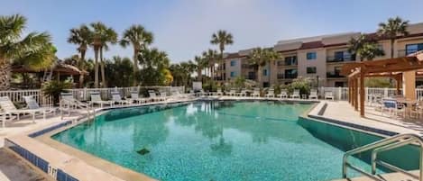 A clear swimming pool with lounge chairs and palm trees, surrounded by a building with multiple balconies.