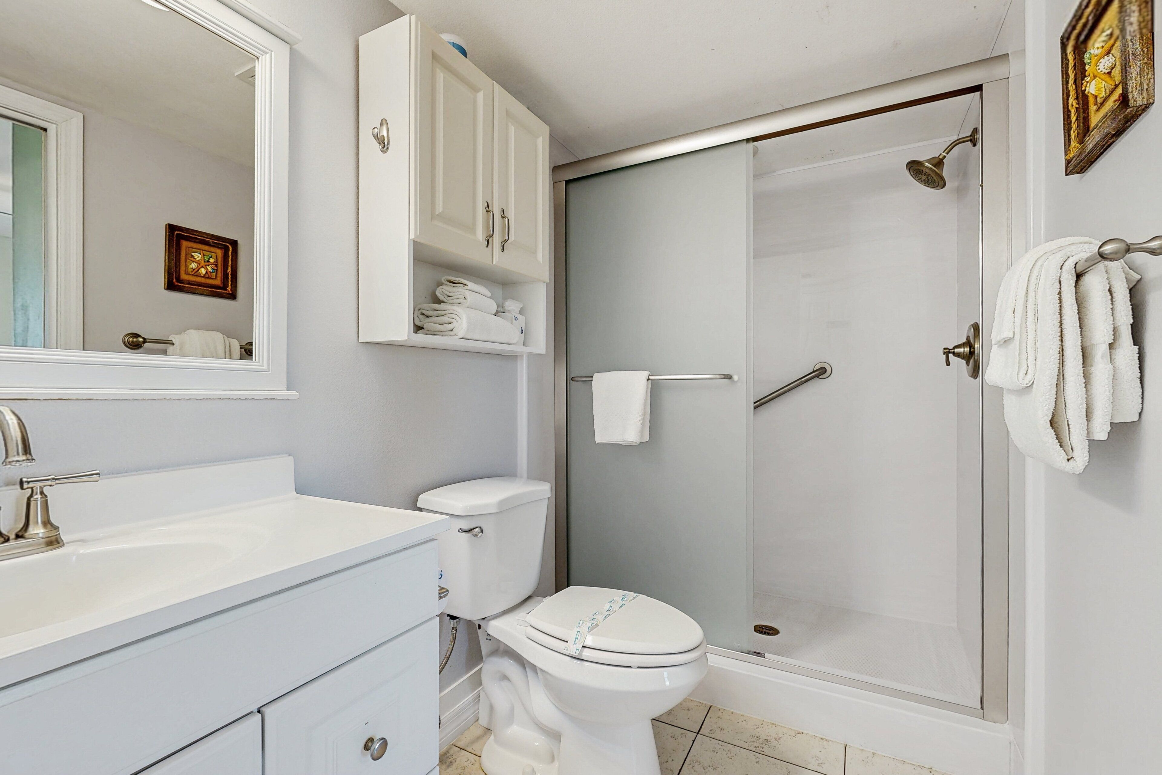 Une salle de bain avec un lavabo, des toilettes et une douche, le tout de couleur blanche. Des serviettes sont accrochées au mur et un tableau encadré est suspendu au-dessus des toilettes.