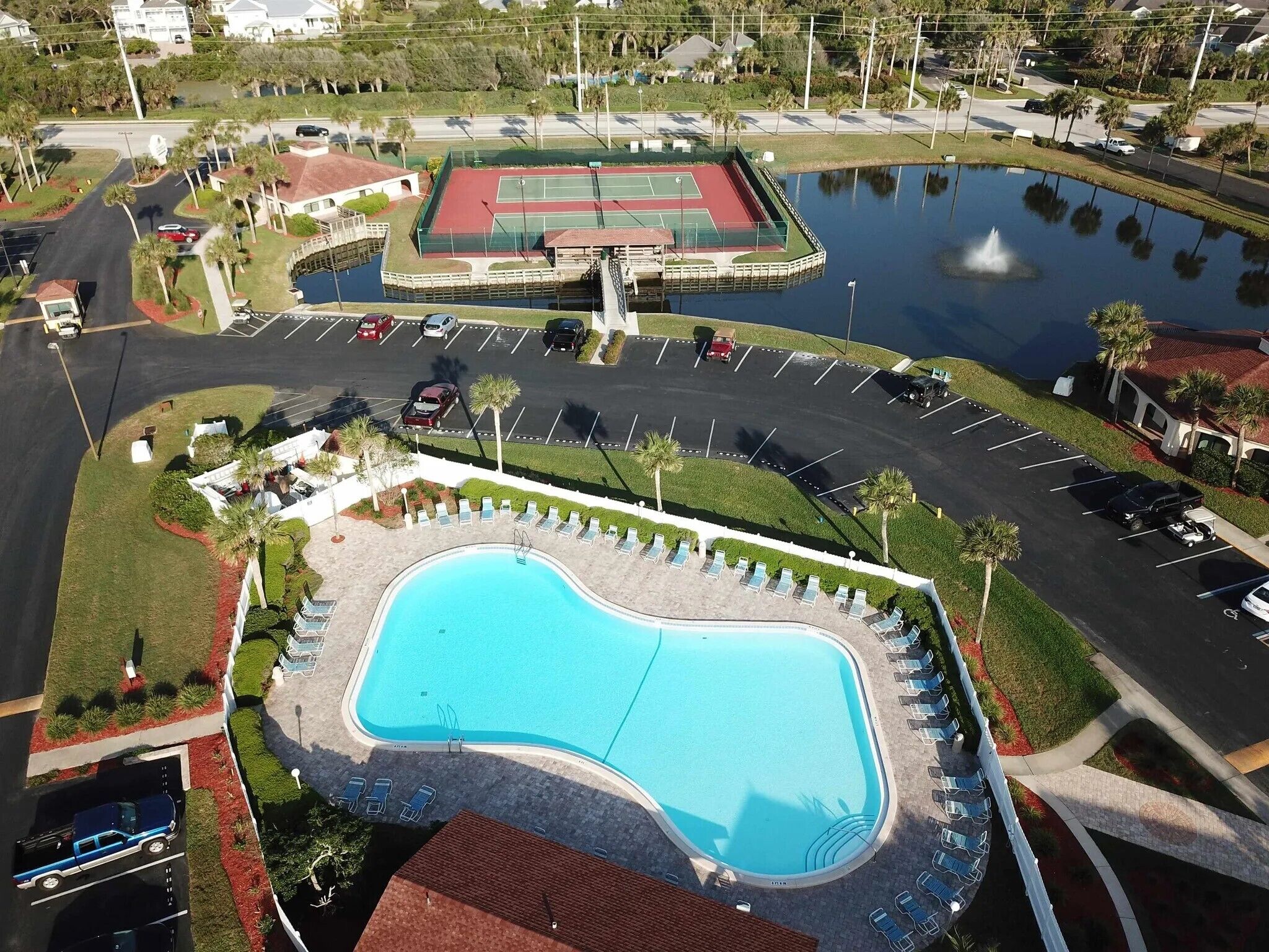 Una piscina con sillones de descanso, cancha de tenis y estacionamiento.