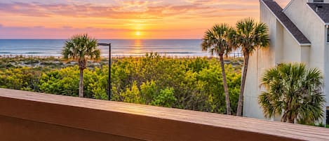 A balcony with a view of the ocean at sunset, palm trees, and a building with a sloped roof.
