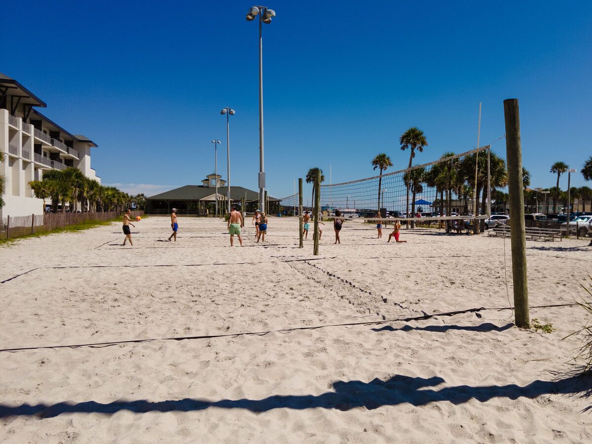 A sandy beach volleyball court with a net, palm trees, and a building in the background.