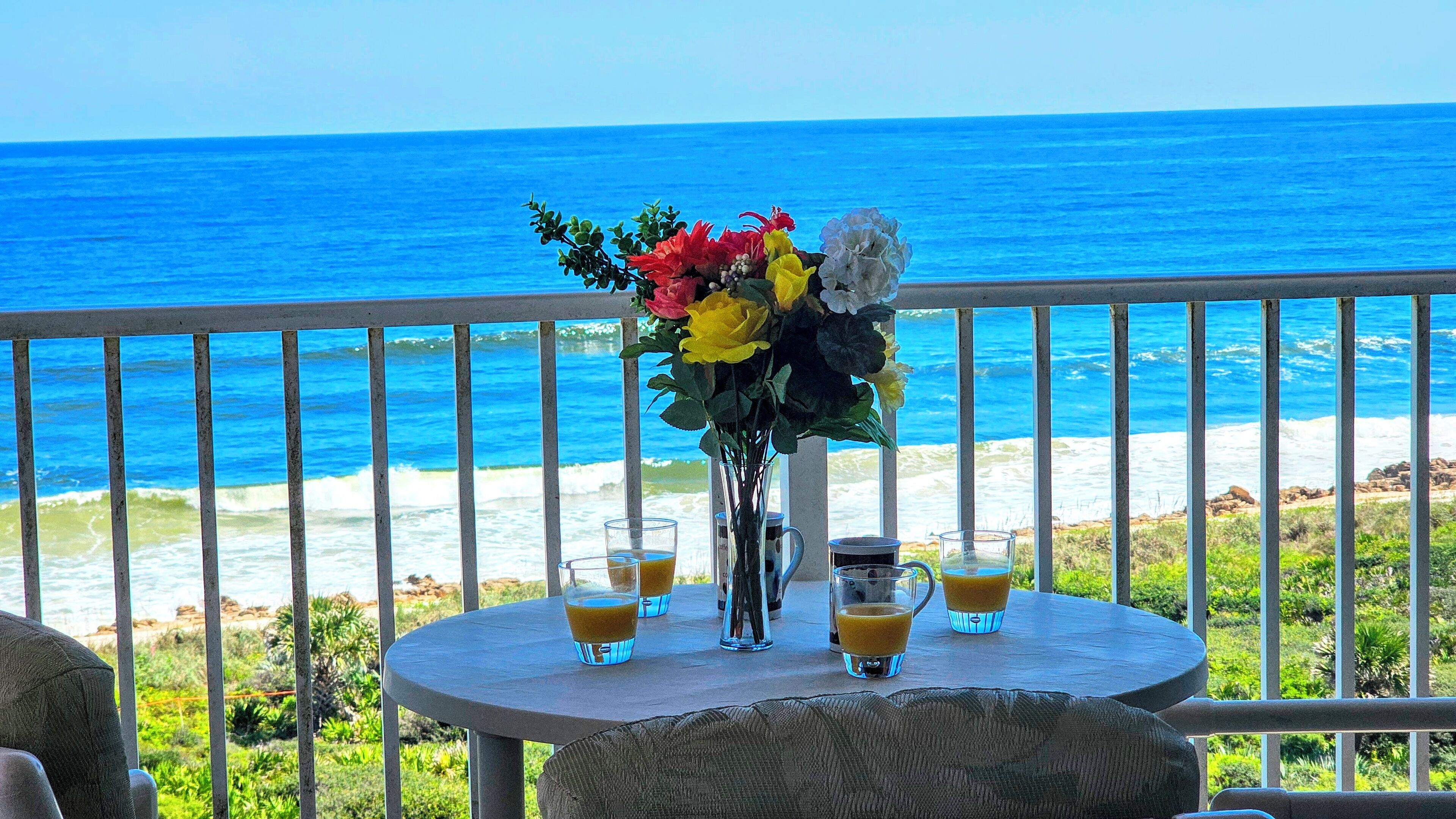 A round table with a vase of flowers and four glasses of orange juice, overlooking a beach with the ocean in the background.
