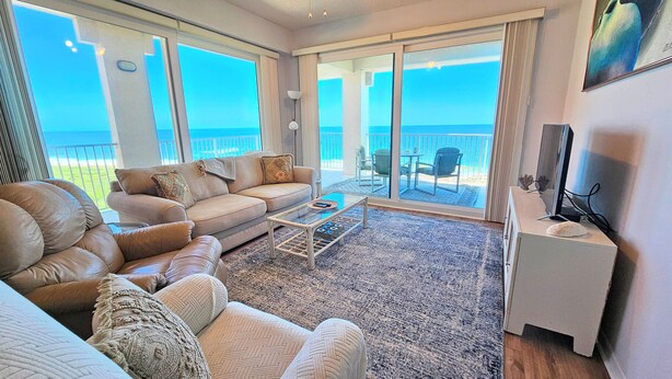 A spacious living room with a large sofa, a coffee table, and a television on a stand. - Surfclub Retreat 6th Floor Penthouse Corner! Wraparound Balcony! Ocean Views! (Palm Coast)