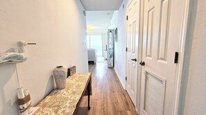 Un pasillo estrecho con suelo de madera, paredes blancas y una puerta a la derecha. Un banco con un cojín estampado está apoyado en la pared izquierda.
