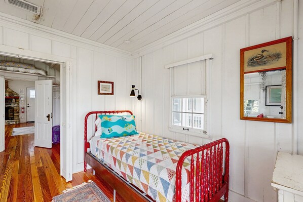 A bedroom with a wooden floor, a bed with a colorful quilt, a red headboard, a window, and a mirror on the wall. - Sunny Oceanfront Home With Balcony, Full Kitchen, Central AC, & Washer/dryer (St. Augustine)