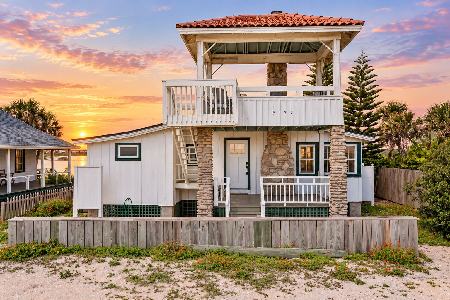 A two-story beachfront house with a balcony, a stone chimney, and a wooden deck.