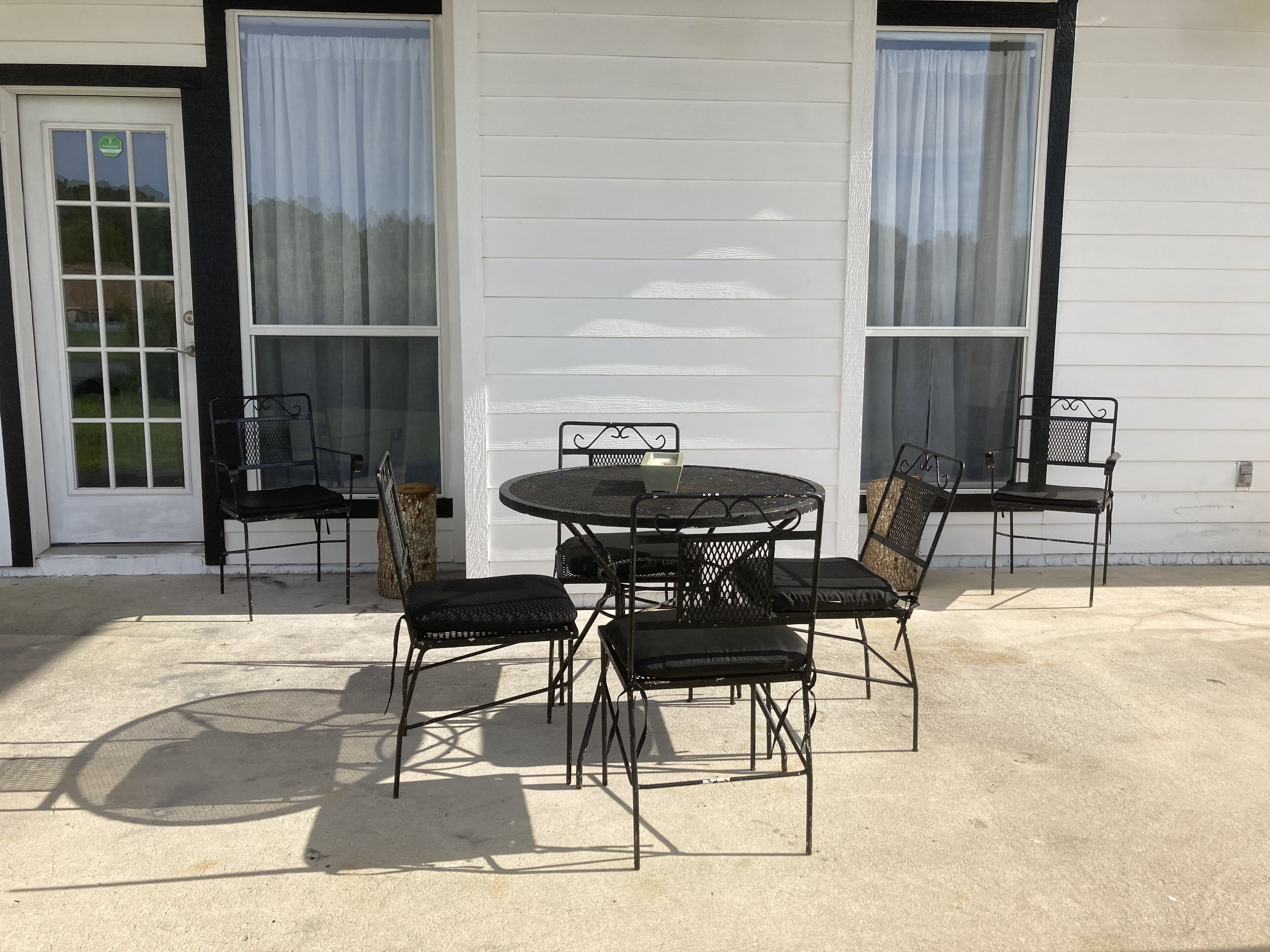 Eine Terrasse im Freien mit einem runden Tisch und vier Stühlen, einem Holzbalken und einem weißen Gebäude mit schwarzem Zierstreifen und Fenstern.