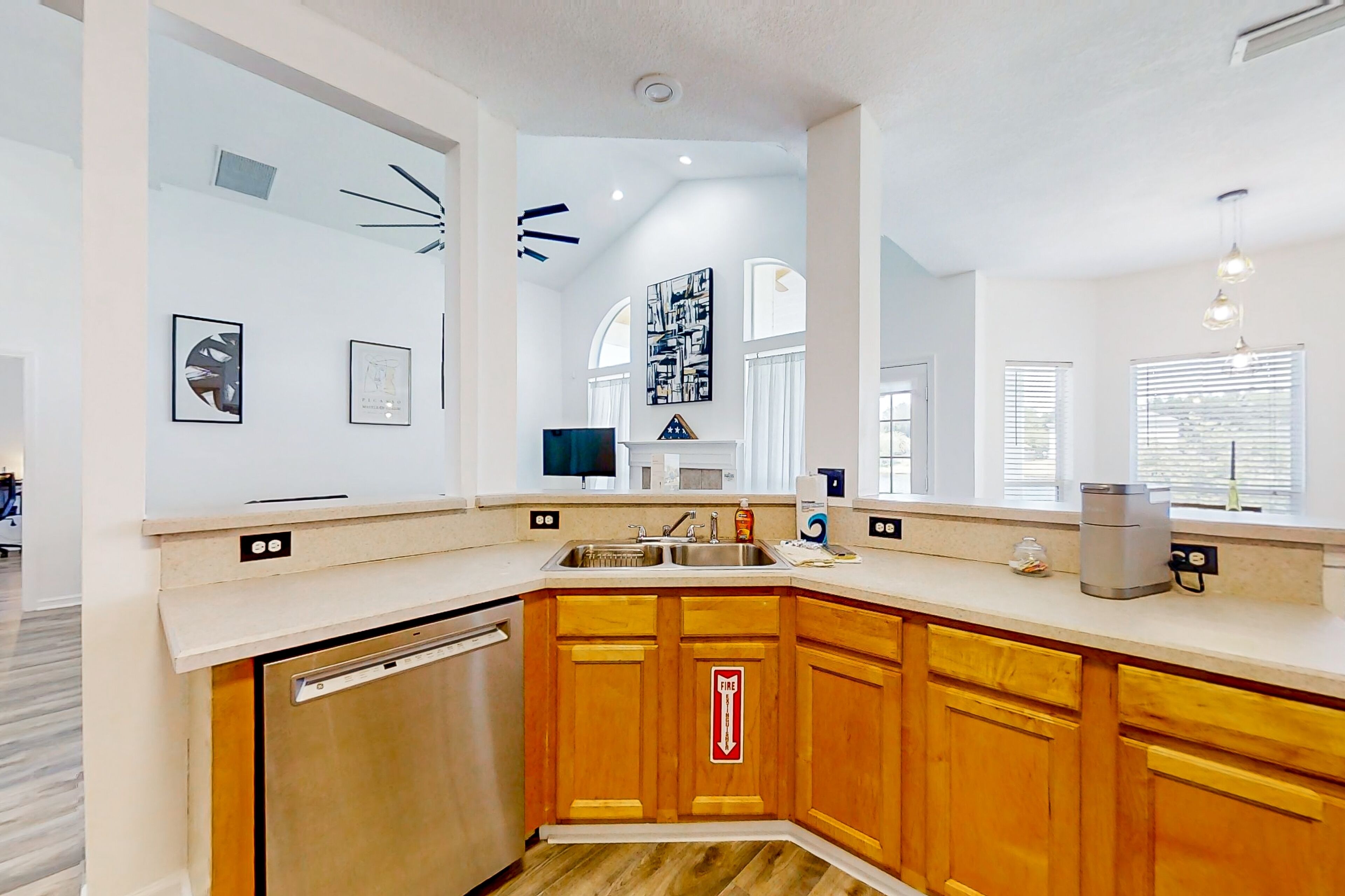 A modern kitchen with wooden cabinets, a dishwasher, and a sink. There are framed pictures on the walls and a ceiling light fixture.