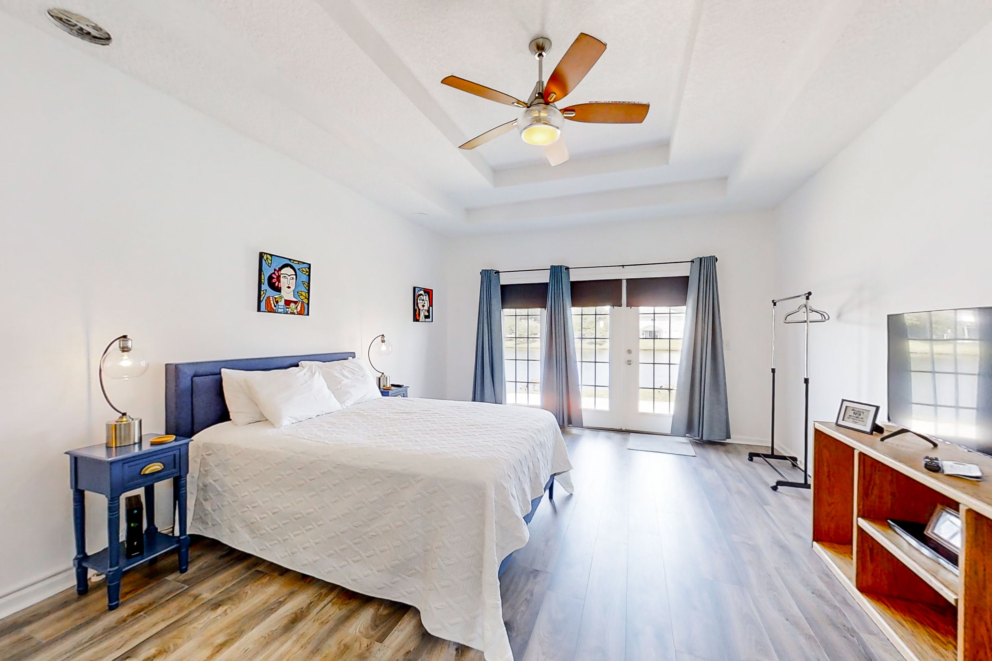Une chambre à coucher avec un lit, un ventilateur de plafond, une table de chevet, un téléviseur et une fenêtre avec des rideaux.
