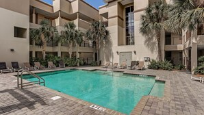 A swimming pool with a 6 ft depth warning, surrounded by lounge chairs and palm trees, with a multi-story building in the background. - Wonderful Home Close to Beach W/shared Pool, Free Wifi, Central AC, W/D (Fernandina Beach)