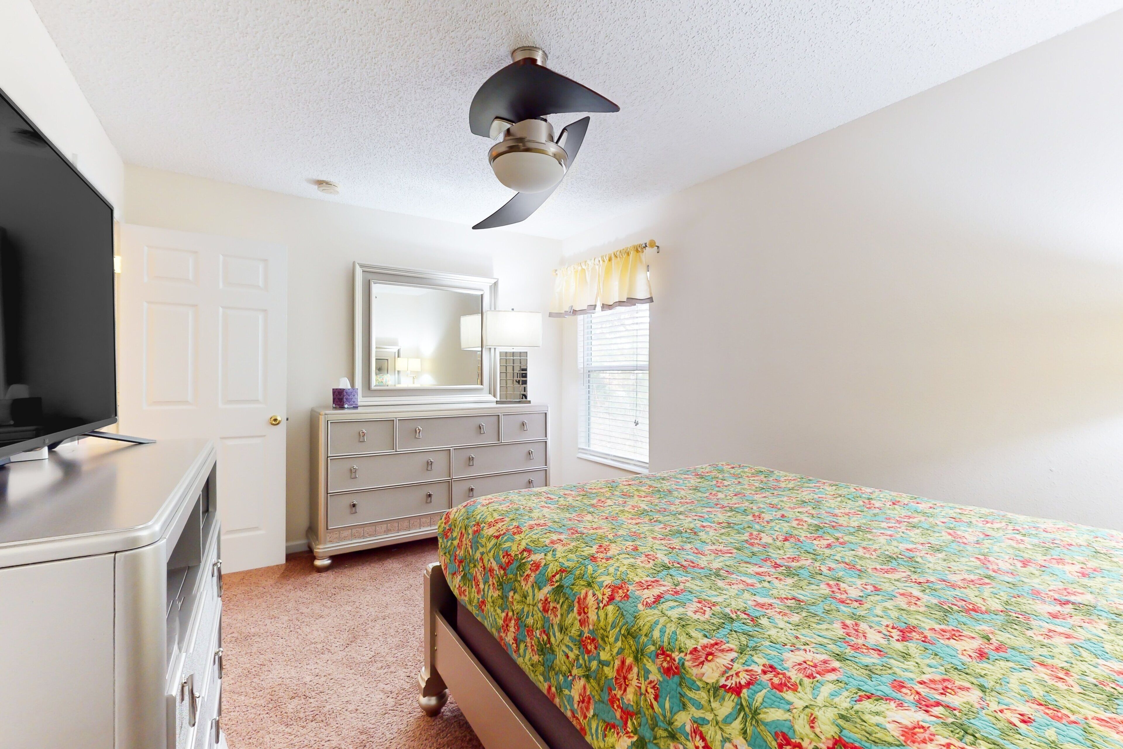 Une chambre  à coucher comprenant un lit, une commode, une coiffeuse, un ventilateur de plafond et une fenêtre avec des rideaux.