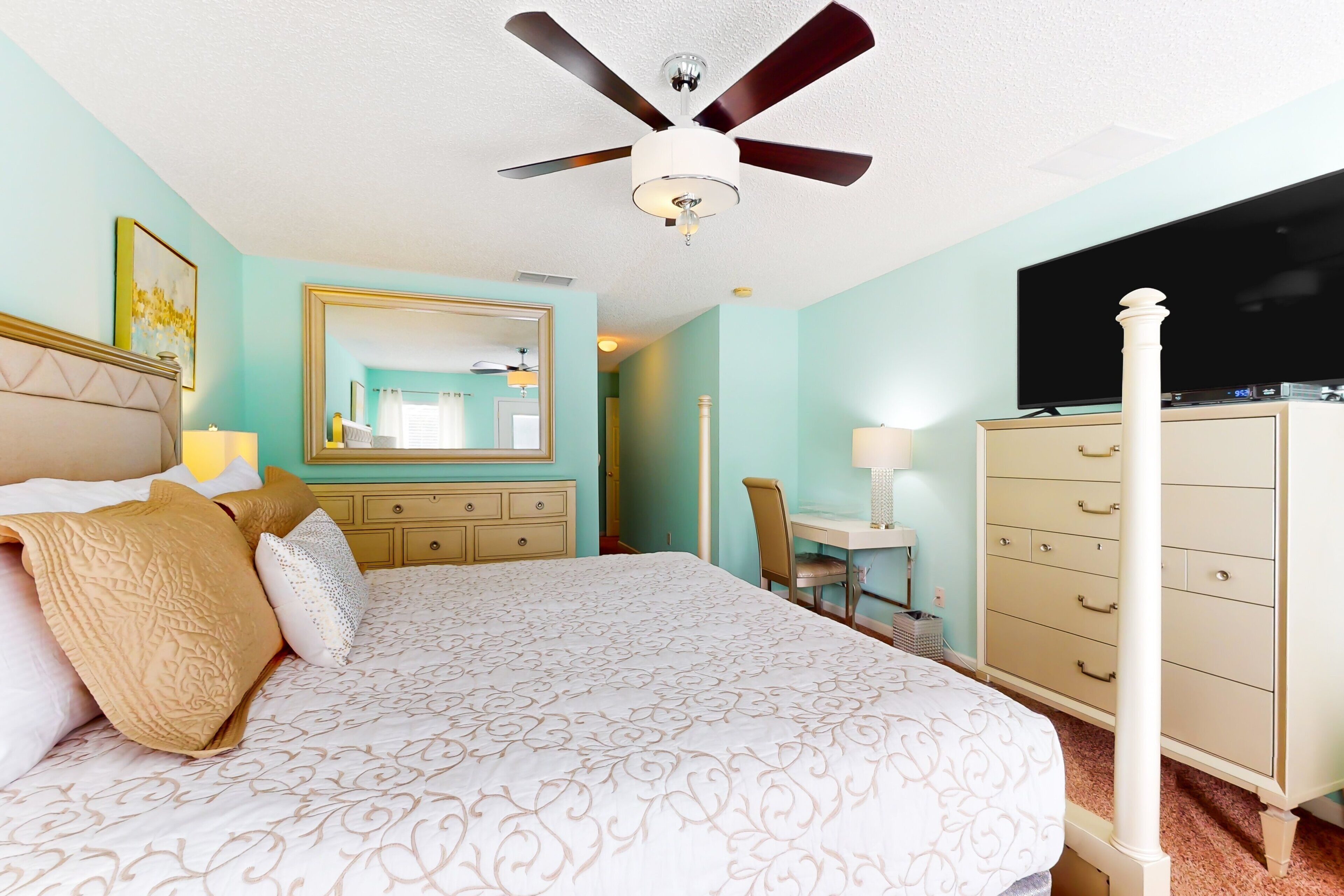 A bedroom with a bed, a dresser, a desk, a television, and a ceiling fan.