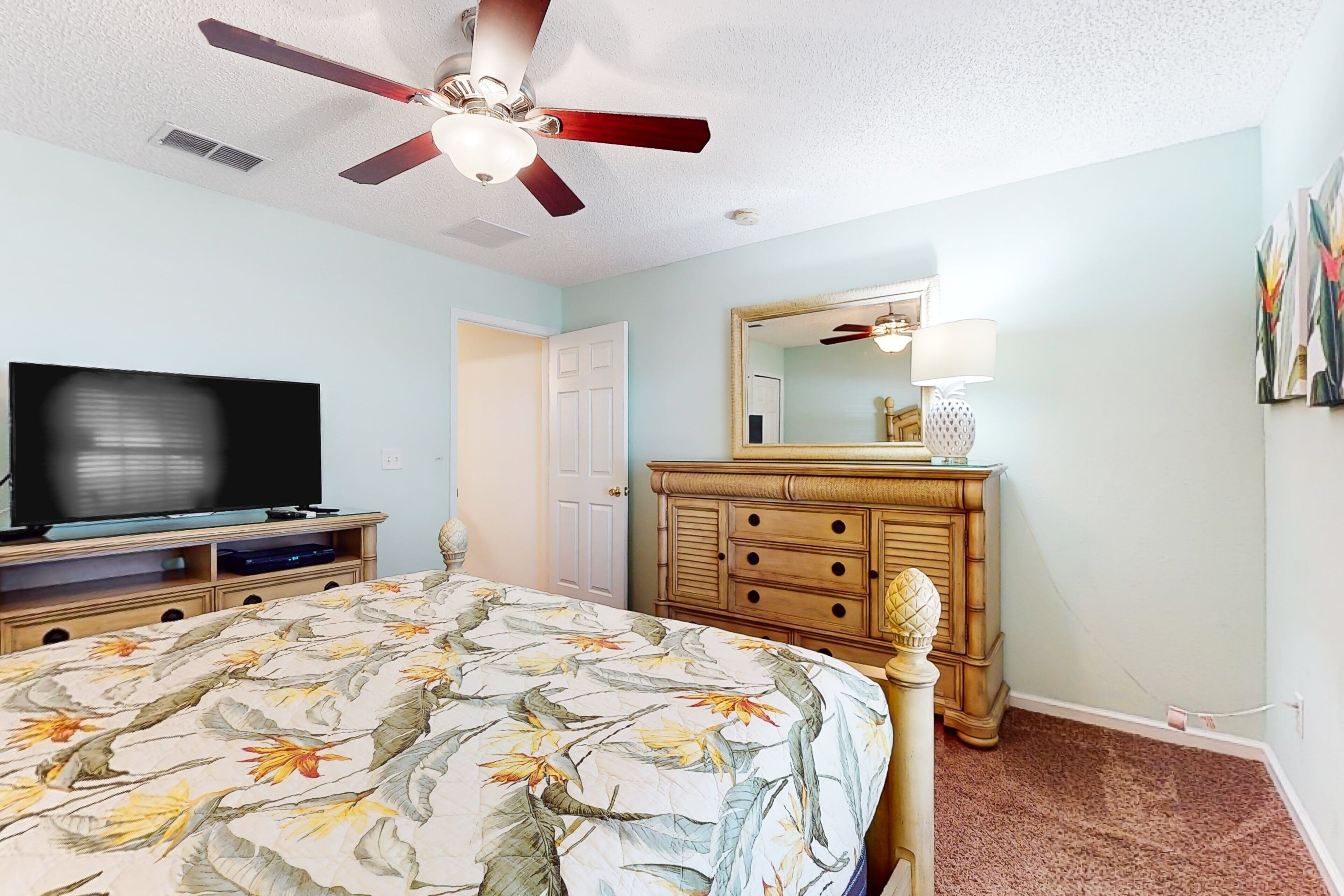 A bedroom with a bed, a television, a ceiling fan, a dresser, and a mirror.