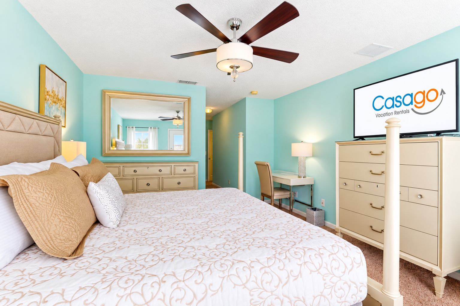 A bedroom with a bed, a desk, a chair, a dresser, a ceiling fan, and a TV displaying 'Casago Vacation Rentals'.