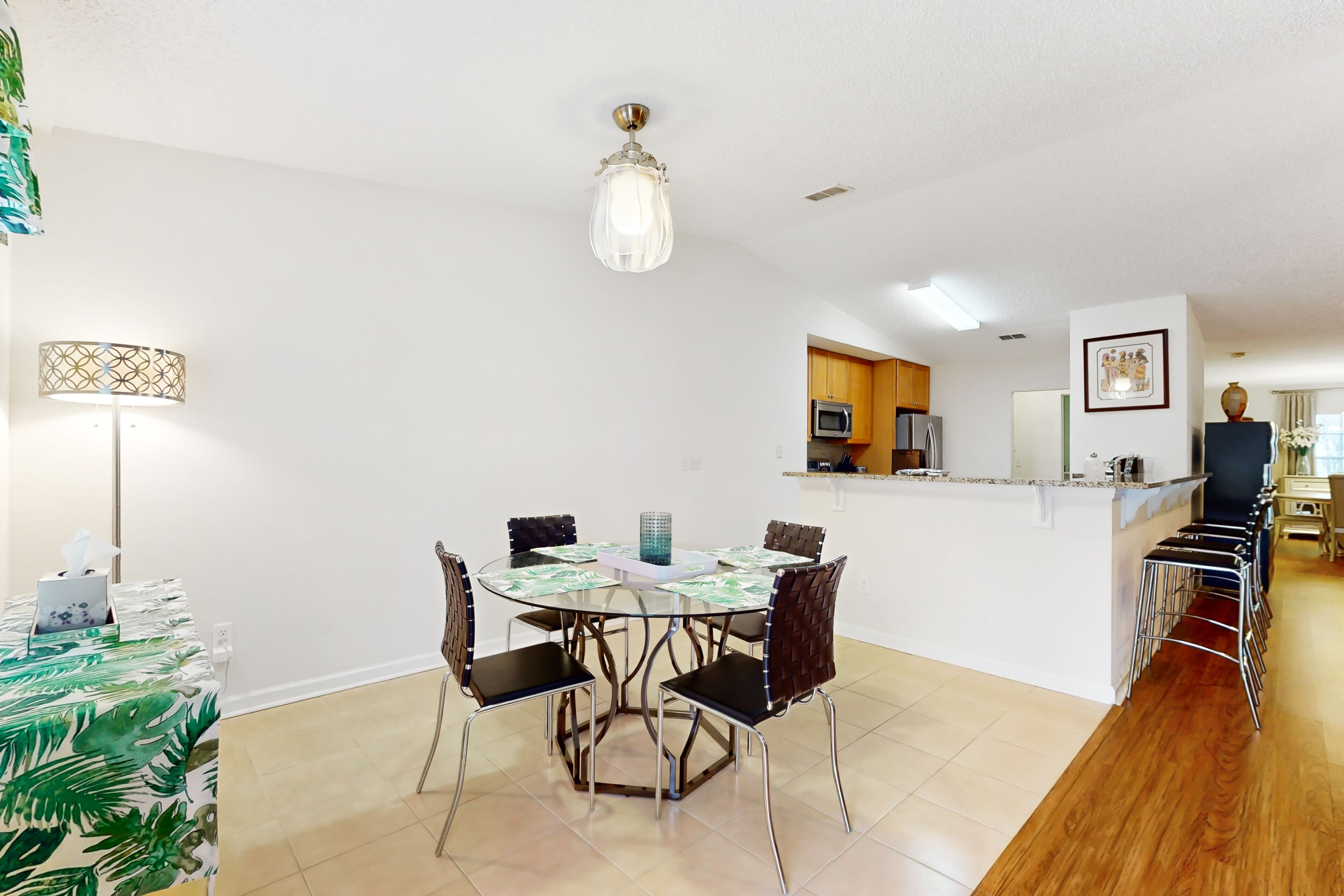 A dining area with a round table, chairs, and a chandelier. There is a kitchen area with cabinets and a microwave.