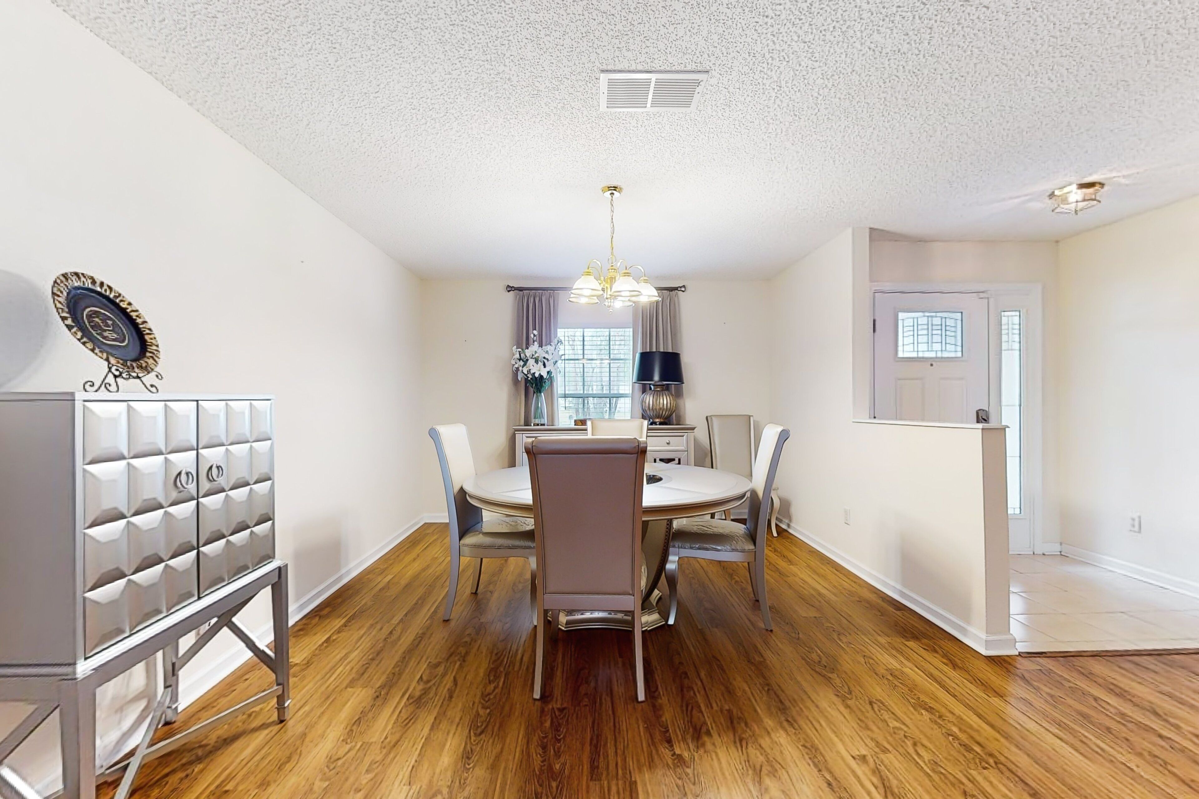 A dining area with a round table, chairs, and a chandelier. There is a cabinet with drawers and a door with a decorative design.