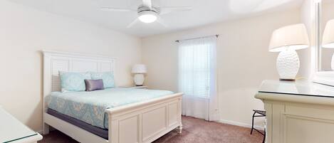 A bedroom with a bed, a ceiling fan, a nightstand with a lamp, a dresser, and a window with curtains.