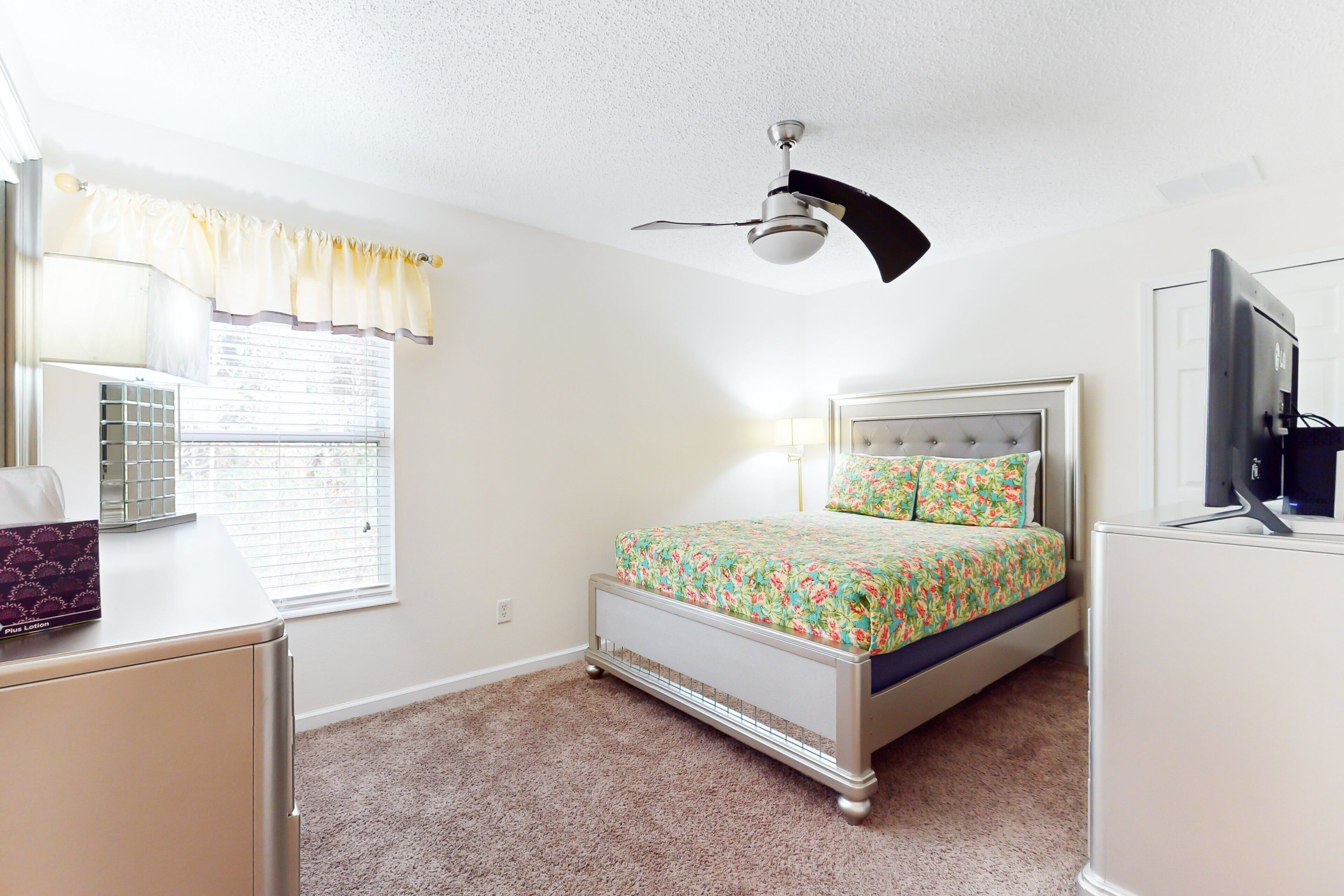 A bedroom with a bed, a ceiling fan, a window with blinds, a dresser, and a television.