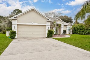 A single-story house with a beige garage door, a front yard with a lawn and a small tree, and a clear sky. - Wonderful Coastal Home With Wifi, Enclosed Backyard, Screened Patio (Palm Coast)