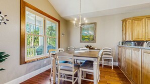 Ein Esszimmer mit Holztisch und -stühlen, einer Chandelierleuchtre und Blick auf Grünpflanzen durch große Fenster.