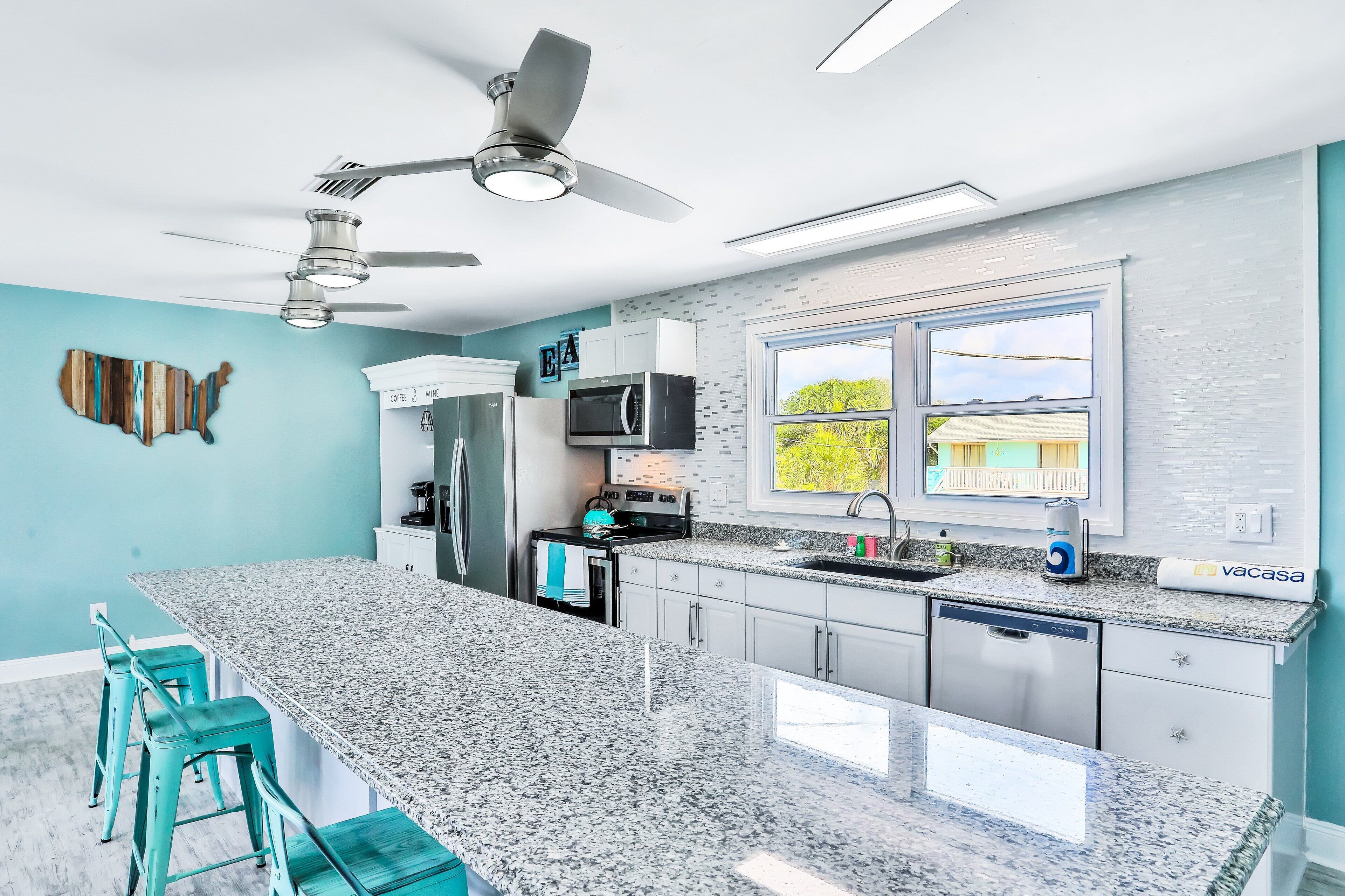 A modern kitchen with granite countertops, stainless steel appliances, teal bar stools, and a large window with a view.